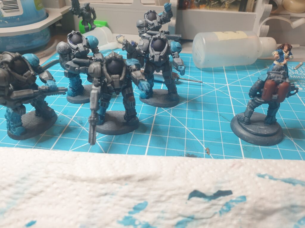 Several miniatures in grey power armour stand on a teal blue craft mat. Each has one shoulder pad, boot and glove painted in bright teal blue.