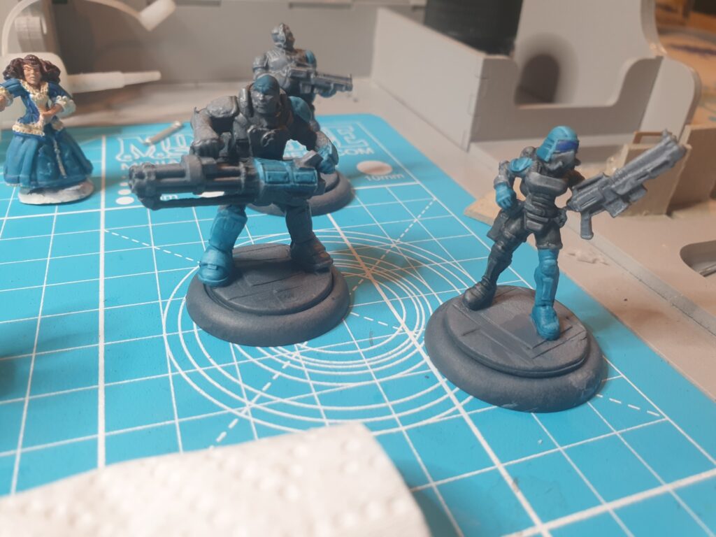 Several partially painted miniatures stand on a cyan craft mat. Each is painted in grey with one bright teal blue shoulder pad and one bright teal blue boot.