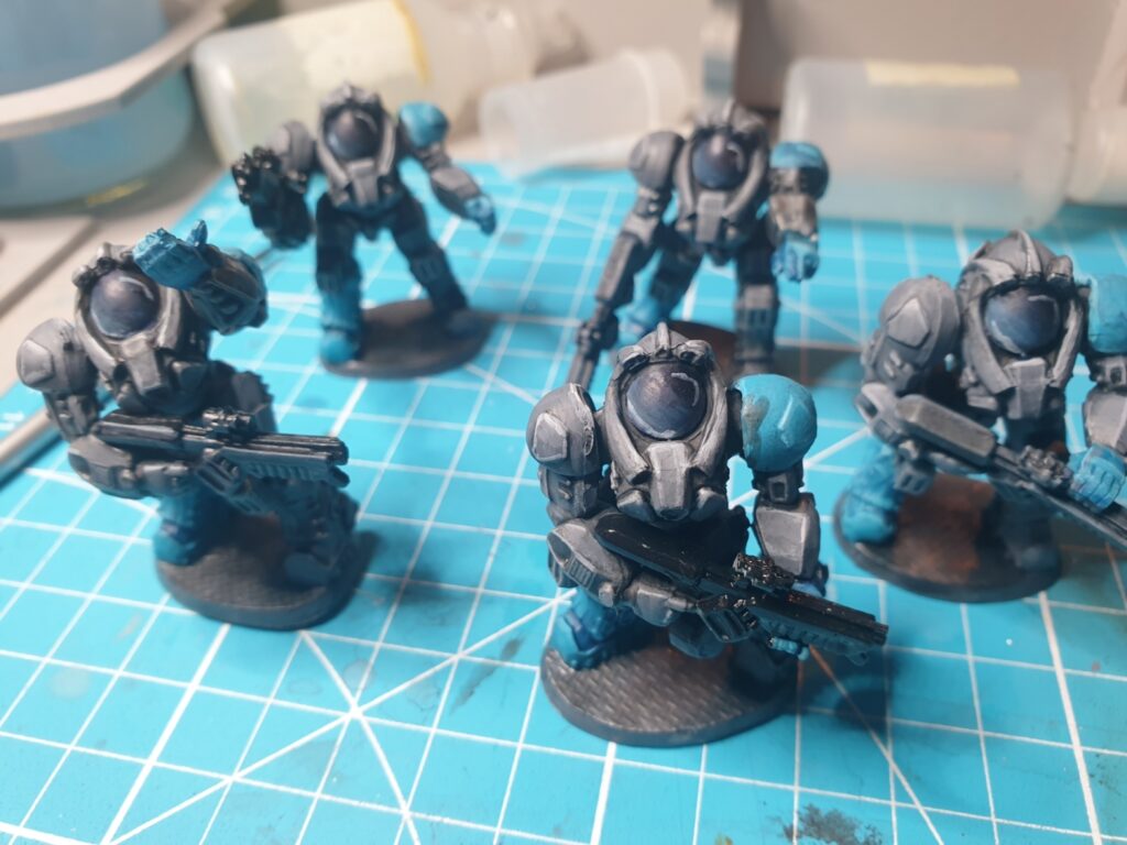 Another photo of five miniatures in grey power armour stand on a teal blue craft mat. Highlights and jewelling has been applied to their glass domed helmets.