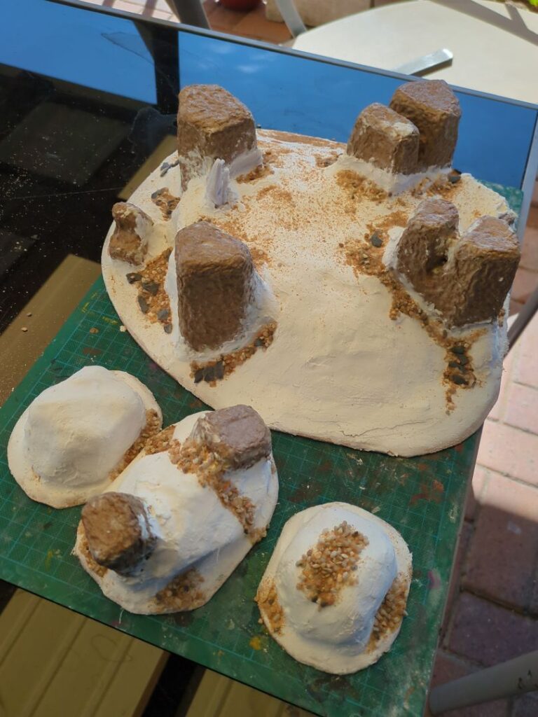 Several plaster hills sit on a green crafting mat. A mixture of sand and rocks have been glue to them to give them a more rocky appearance and texture.