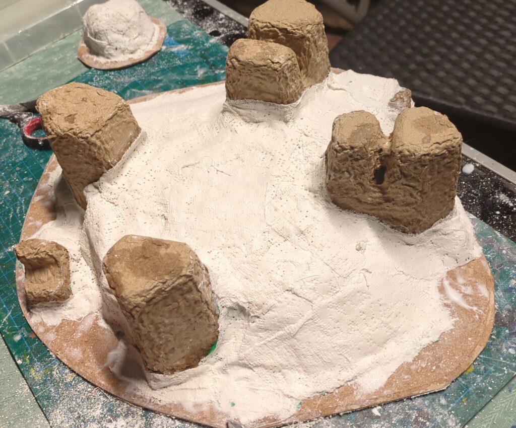 Some cardboard packaging attached to a simple MDF base. It has been covered in white plaster to create a hill.
