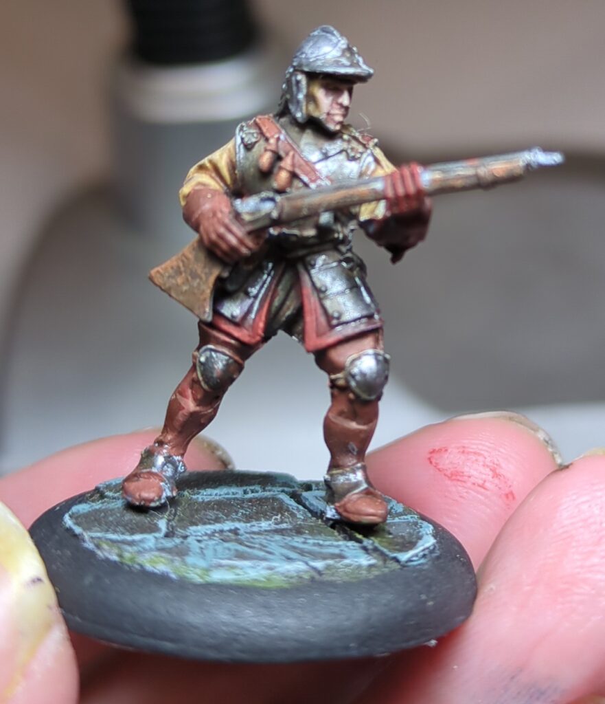A painted figurine of a Hell Dorado Arquebuser stands, firearm at the ready wearing his uniform and armour.