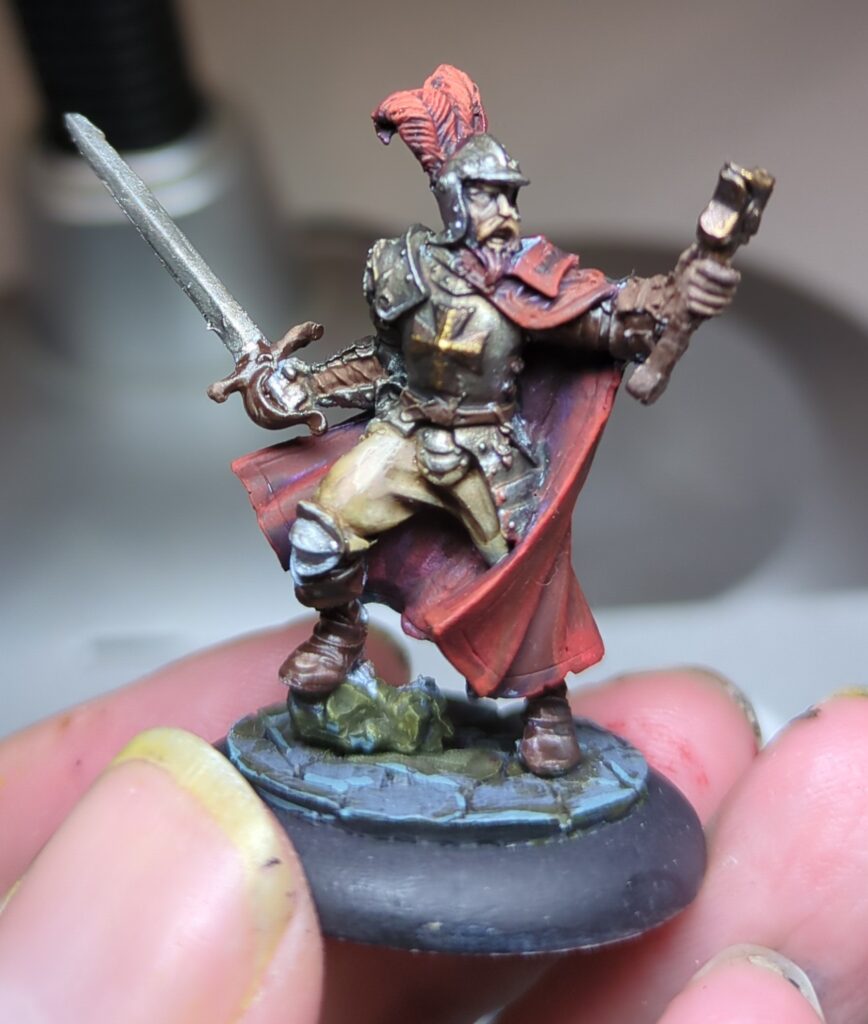 A small hand-painted figurine of Francisco Vargas, a Spanish-style captain from the Hell Dorado Westerners range. His red cloak is flowing around him and a gold cross is emblazoned on his breastplate. He stands holding a holy artifact of some sort in one hand and his sword in the other.