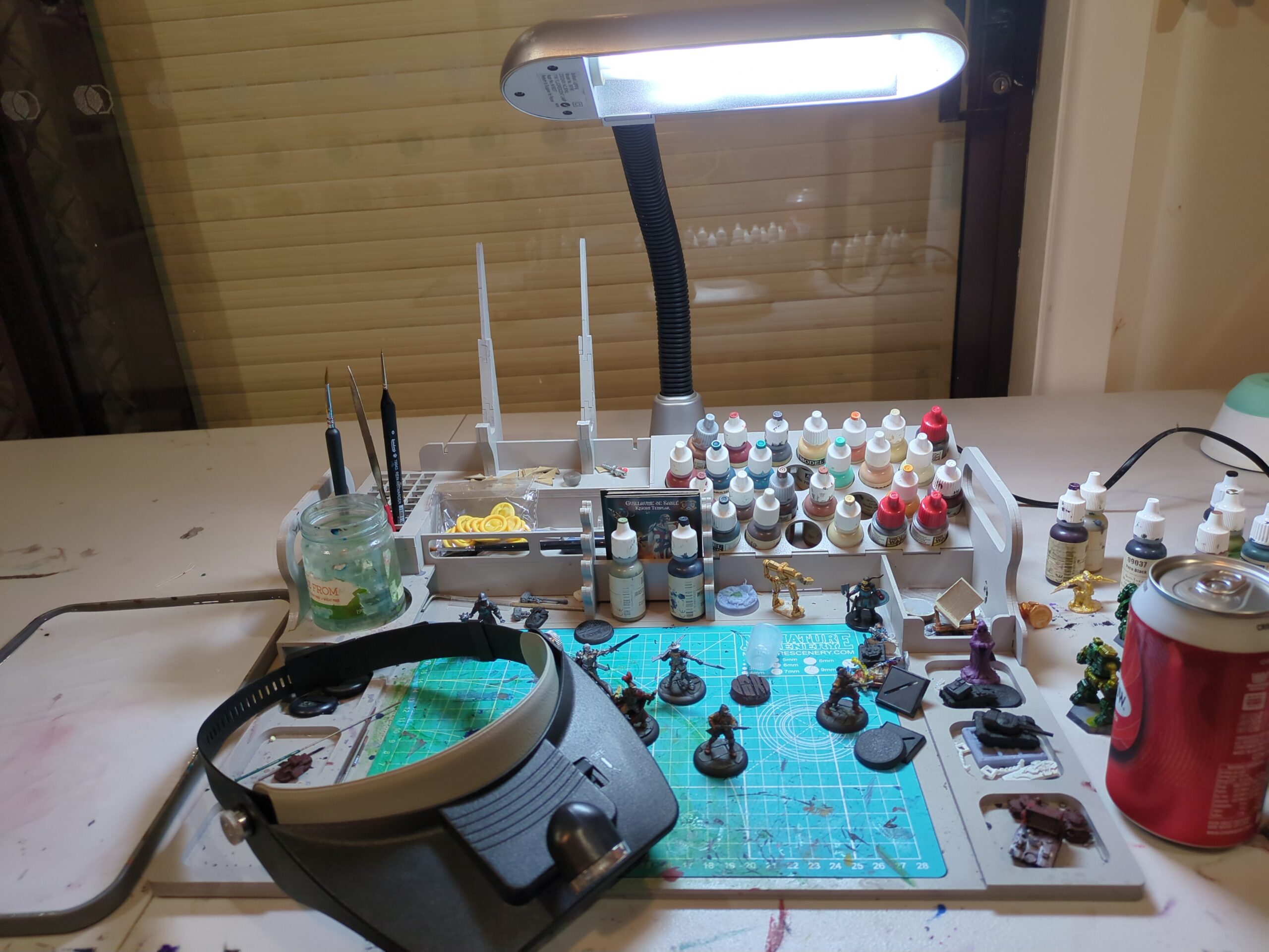 A hobby station surrounded by a wet palette, magnifiers, paints, a light and of course - a cold drink.