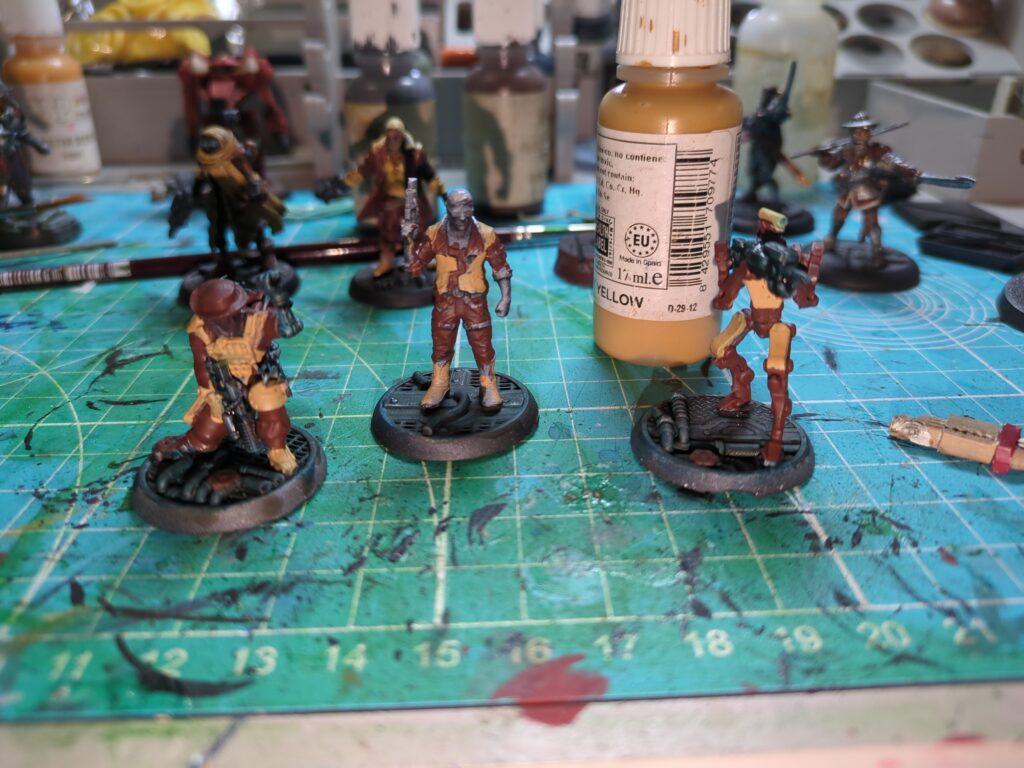 Several figurines, most in various stages of being painted with a red and yellow paint scheme sit on a teal painting mat. One completed figurine painted in the same scheme is also present to act as a reference.