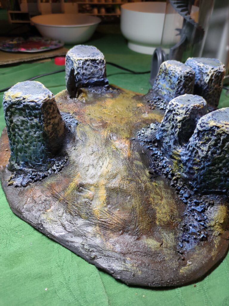 A large terrain piece sits on a table, clearly showing the slope of a hill that rises up towards the centre of four large monoliths.