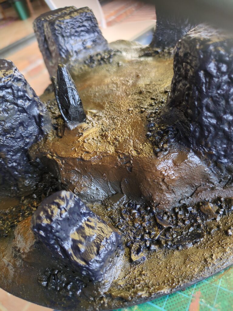A painted plaster hill with rock monoliths protruding from the earth. So far, only a base coat has been applied, so the hills are a dark flat brown and the monoliths are a dark blue-grey.