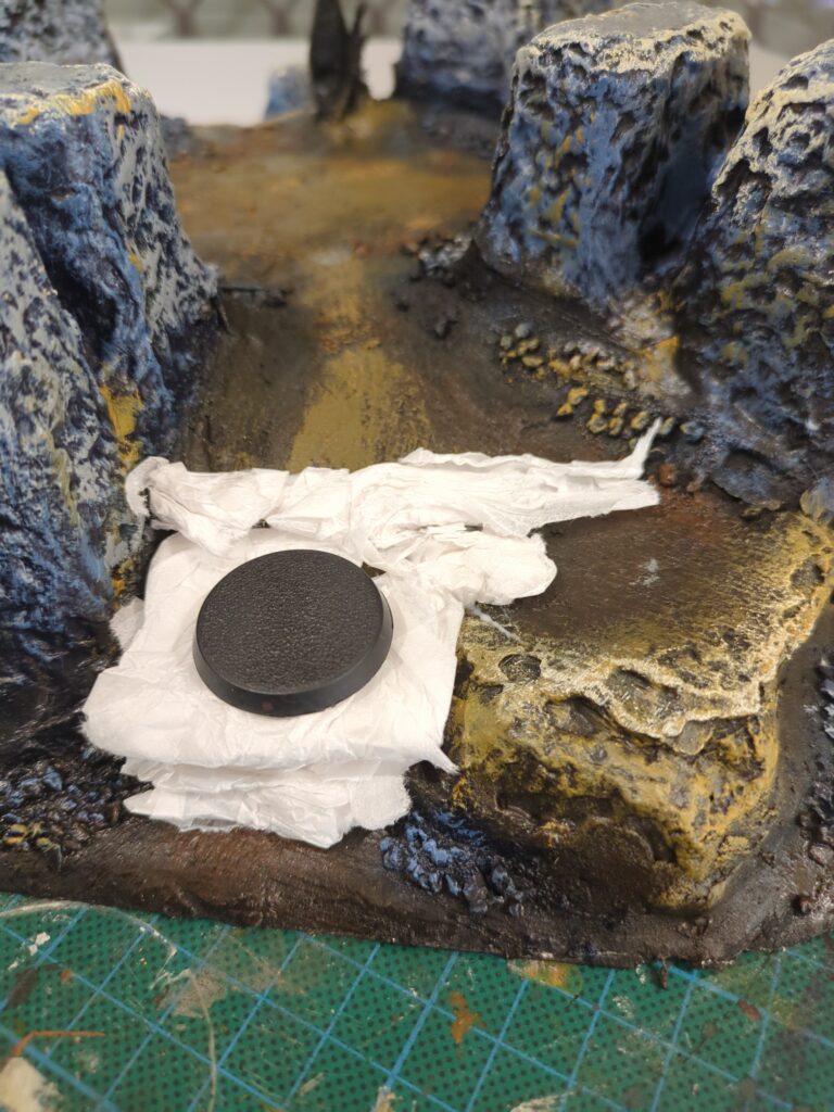 A close up of a plaster mountain with tissue paper added to create the foundation of a new small ledge. A round plastic base sits atop it, testing the size.
