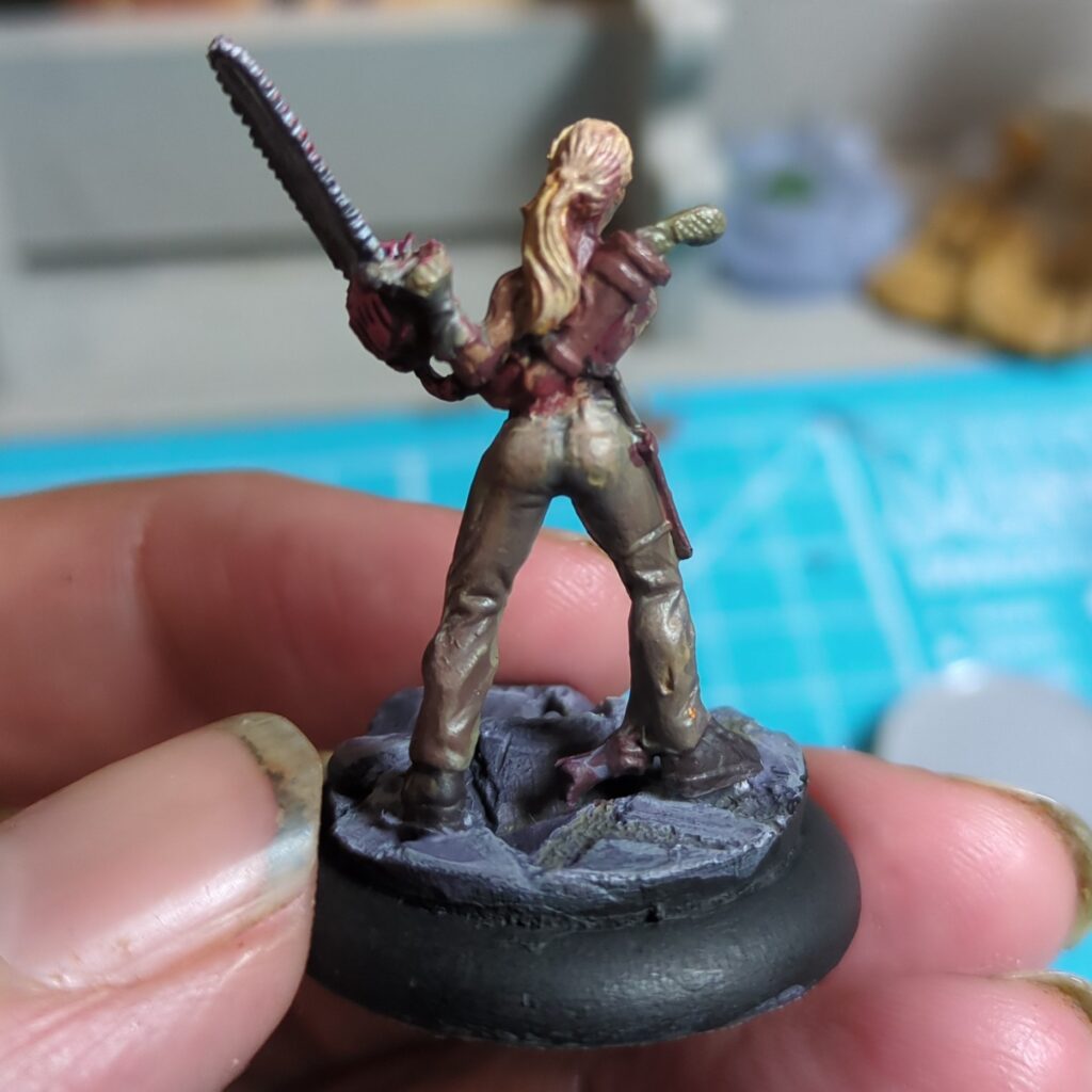 A view of the back of figurine of a woman. She is standing in a ruined street and brandishing a chainsaw. She is wearing a red top and khaki pants, and has a large sheathed knife on her back.