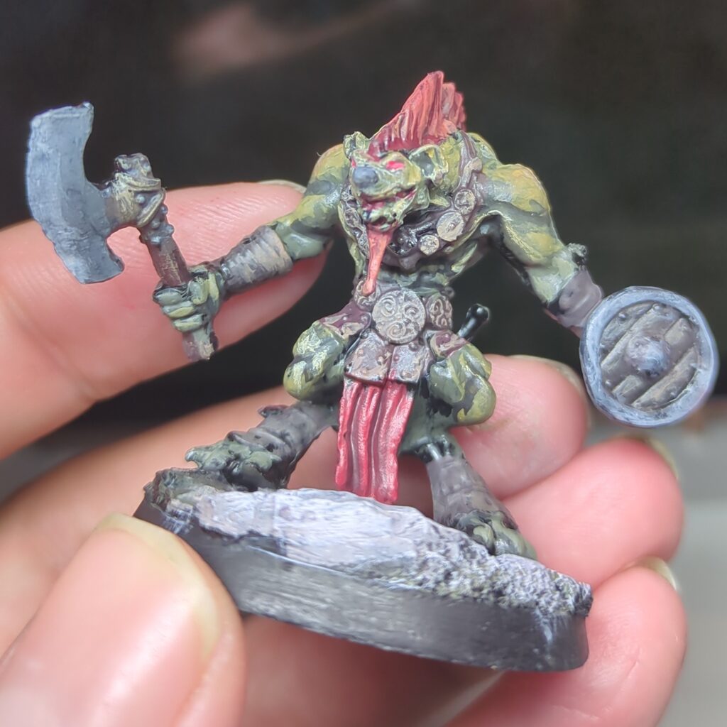 A gnoll with greenish yellow skin. He has red hair and wears a mix of leather and red cloth. He stands brandishing an axe and shield.