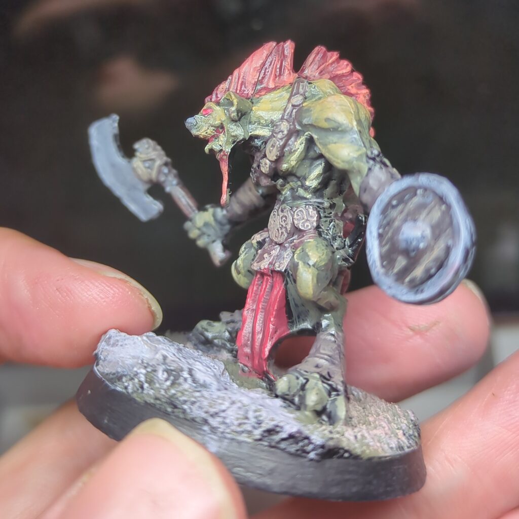 Side view of a gnoll with greenish yellow skin showing off his bright red Mohawk and the details of his face. He has a small black nose and his jaw is set in an angry snarl.