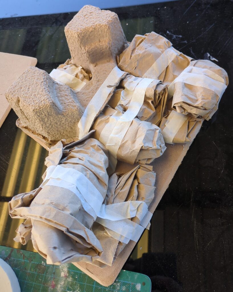 Several bits of balled up paper and part of some cardboard packaging are haphazardly taped down on an MDF base.