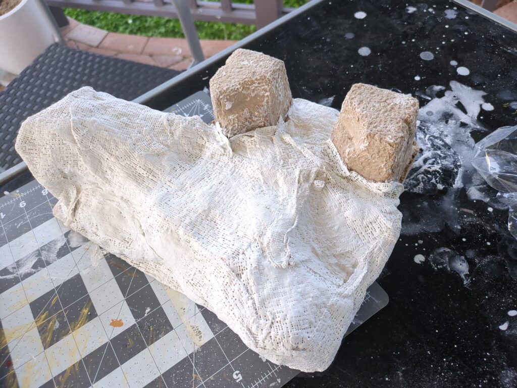 Plaster bandages have been laid down to create a crude mountain form. Some of the packaging material can be seen protruding from the bandages, vaguely resembling stone ruins
