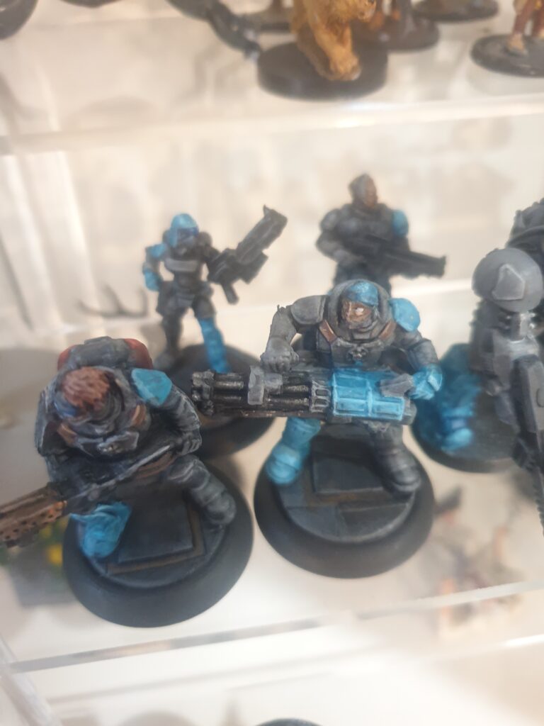 Four painted soldiers stand on an acrylic shelf. They wear body armour and have an assortment of weapons (one flamer, one gattling cannon and two rifles). All of them have one bright teal shoulderpad and one bright teal boot.
