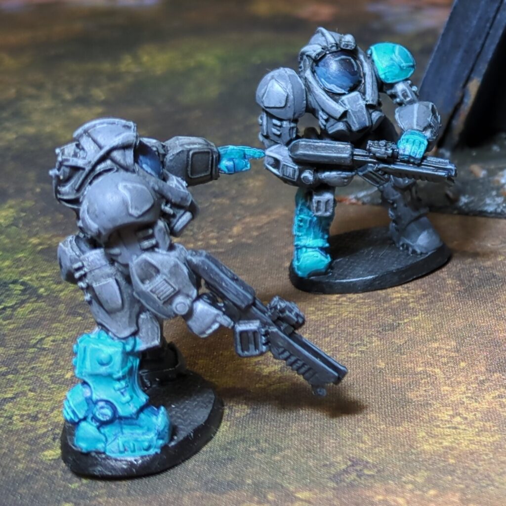 Two troopers in dark grey power armour stand near a bunker. On each one boot and the opposite shoulder pad are painted bright teal.