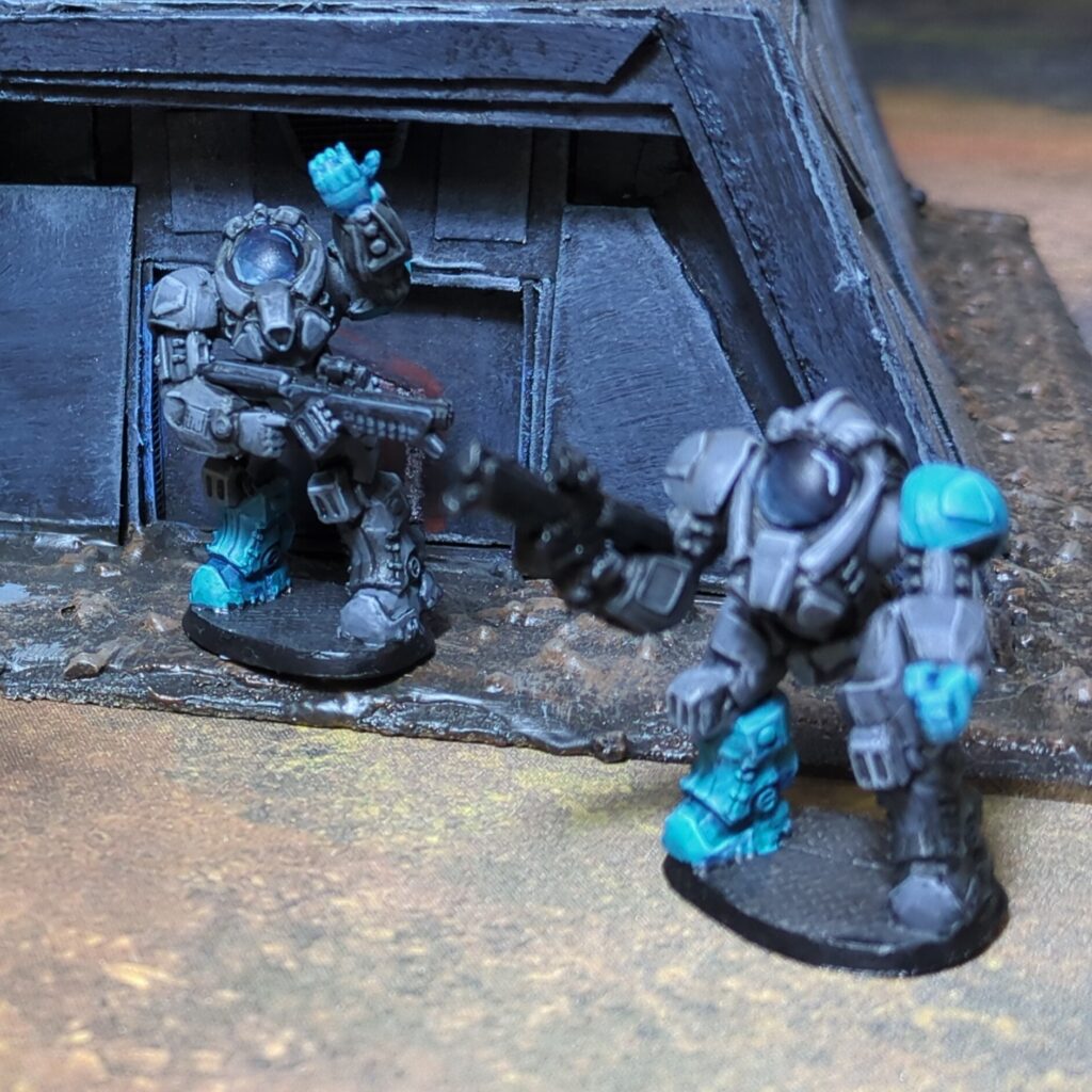 Two troopers in dark grey power armour stand in front of a dark grey bunker. Both hold dark black rifles and have a shoulder pad, boot and glove that are bright teal.