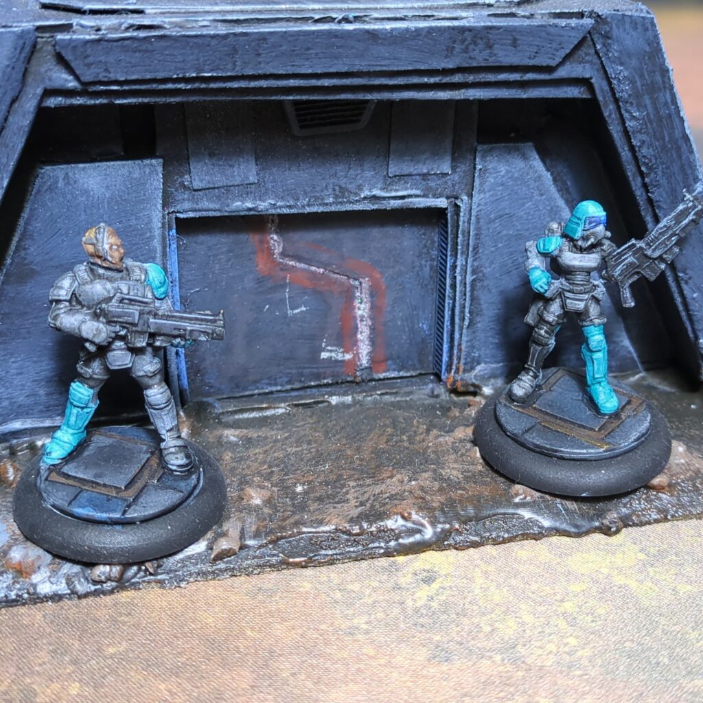 Two soldiers in grey body armour stand in front of a grey bunker. Both have one teal shoulderpad and one teal boot. One also wears a teal and grey full-face helmet, with the other wearing a communication headset.
