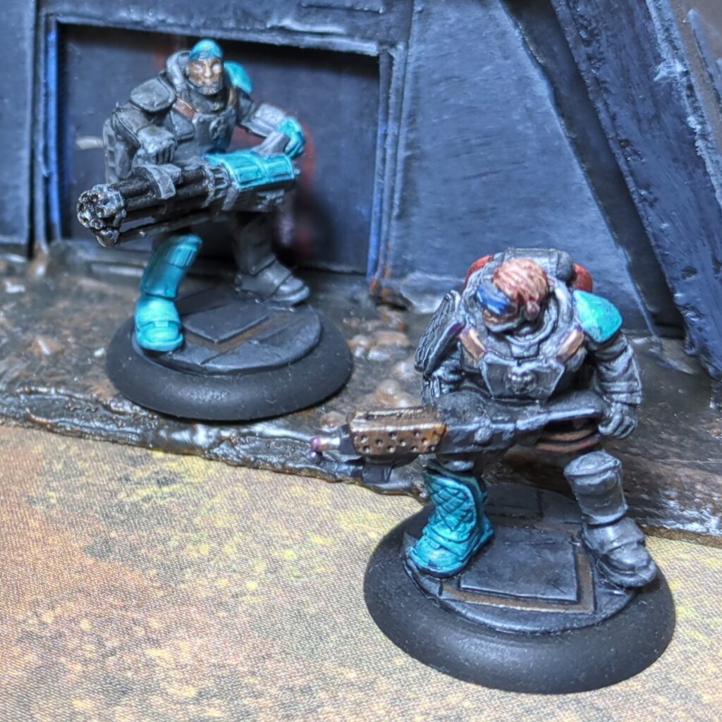 Two soldiers in grey body armour stand in front of a dark grey bunker. Each has one teal boot and one teal shoulderpad, with one also sporting a teal bandana. One wields a gattling gun and the other wields a flamer.