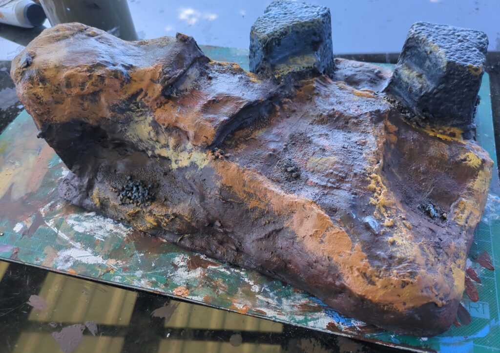 Another piece of terrain sits on a green craft mat. We can see an ascent leading up to a large overhang. At the bottom of the ascent, two large stone blocks flank a small pass.