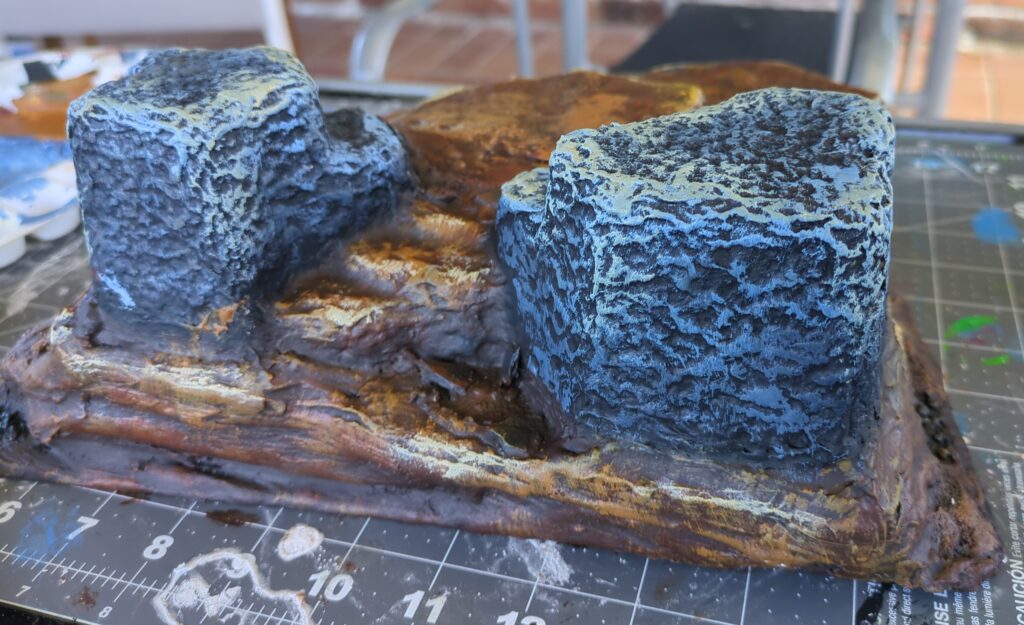 A completed terrain piece sits on a rubber craft mat. Two stone blocks flank a thin set of steps leading up a mountainside.