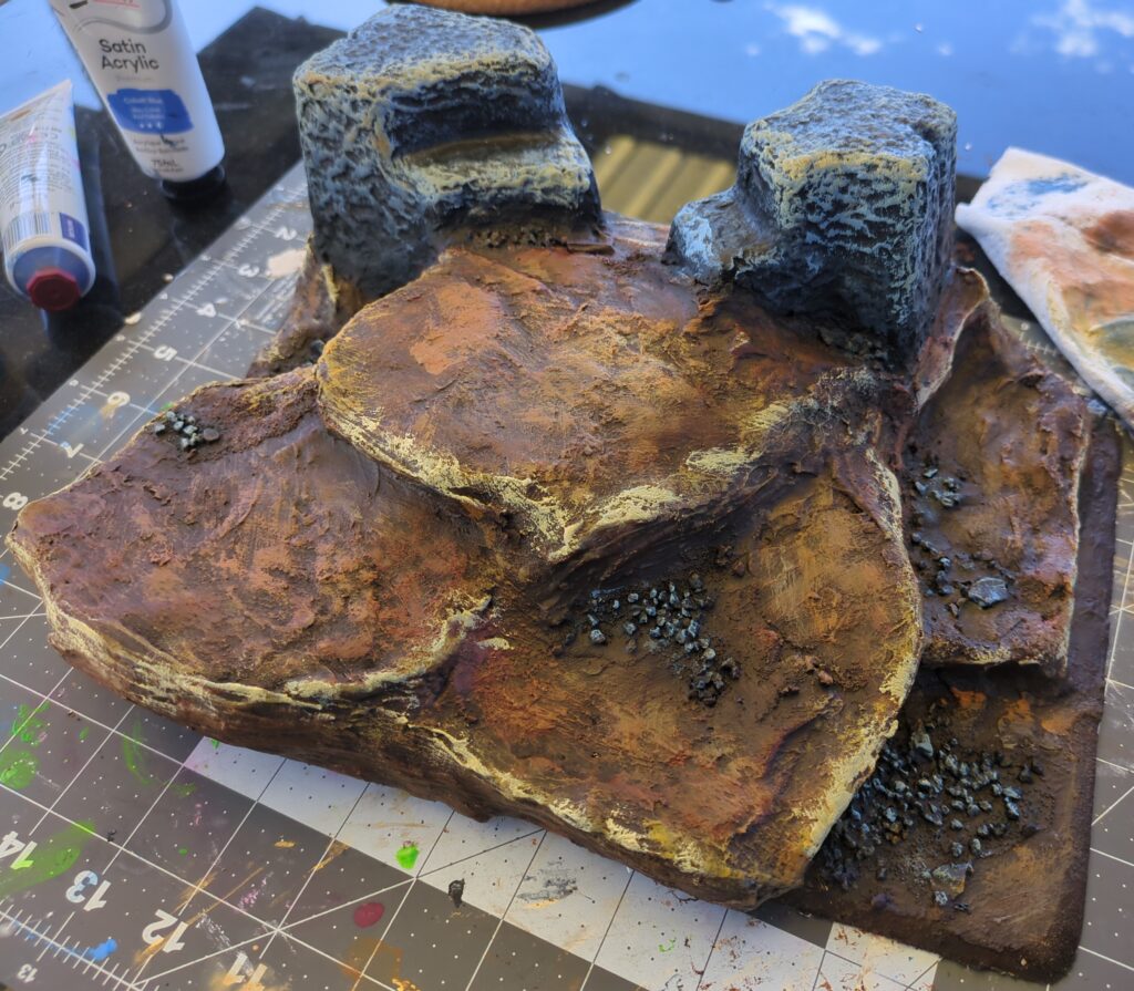 A completed piece of terrain sits on a rubber craft mat. Several ledges can be seen leading up to two large stone blocks with a thin pass between them.